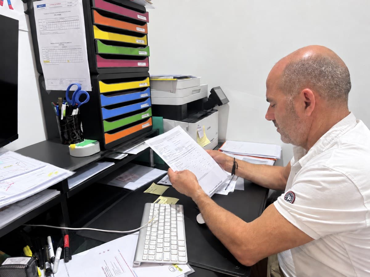 Le père de Robin à son bureau, une commande de chantier à la main, au milieu des papiers.